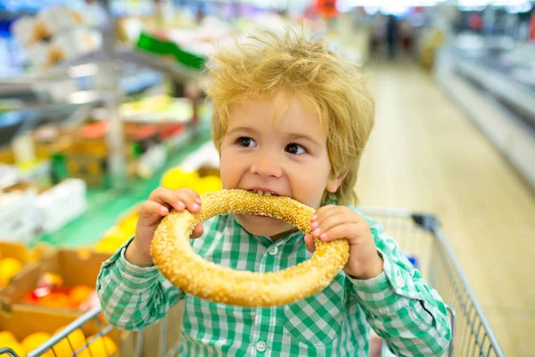 子供はスーパーやパン屋でパンを食べます。子供のためのおいしいペストリー。炭水化物と軽食。ごまのベーグル。ショッピングカートバスケットのスーパーマーケットへの旅行。歯を噛むために. — ストック写真