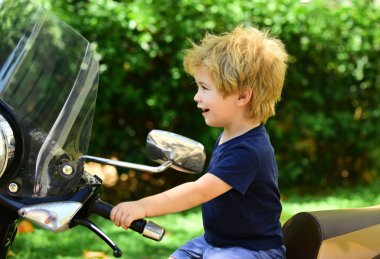 Küçük motosikletçi motosiklet ve gülümsüyor oturur. Motosiklet saplı sarışın çocuk. Yeşil doğal arka plan üzerinde gülümseyen mutlu çocuk. Temiz havada aile tatilleri.