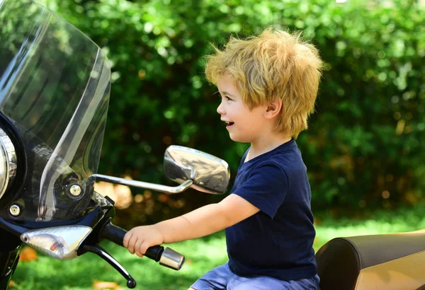 Küçük motosikletçi motosiklet ve gülümsüyor oturur. Motosiklet saplı sarışın çocuk. Yeşil doğal arka plan üzerinde gülümseyen mutlu çocuk. Temiz havada aile tatilleri.