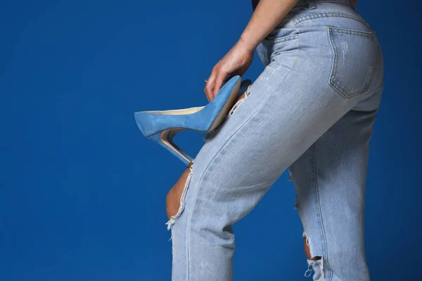 Jeans with holes. New trend, blue color. Beautiful female feet, the girl holds in her hand blue high-heeled shoes. Shoes and clothes, fashion. Style for summer