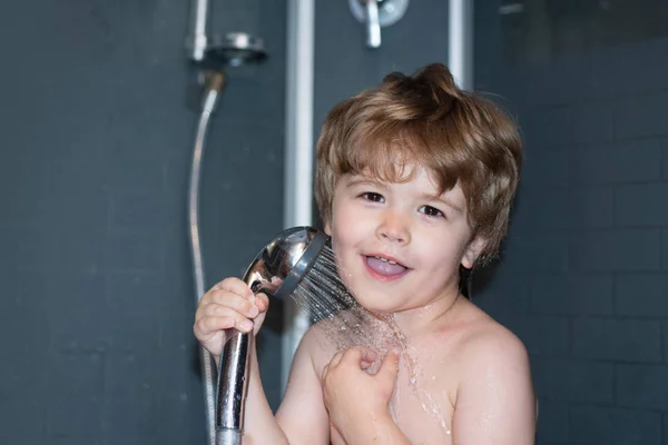 Shower. One beautiful happy laughing baby boy with wet blonde hair in shower. Standing under water indoor looking away on grey plumbing background. Happy time in bath.