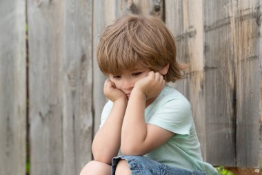 Üzgün çocuk. Bir çocuk ahşap bir çitin yanında oturuyor. Okul öncesi ve sonbahar. Üzücü düşünceler. Hayal kırıklığı.