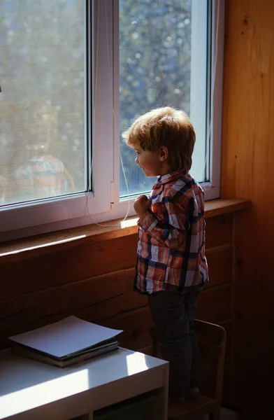 El niño mira por la ventana con curiosidad. La luz del sol entra en la habitación a través de las ventanas. Grandes ventanas de plástico blanco llenas de luz solar. Lindo niño disfrutar del sol en casa a través de ventanas . — Foto de Stock