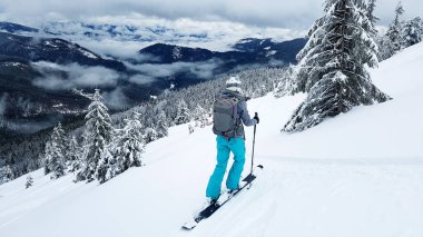 Genç kayakçı kayaklarıyla çakıllı bir yamaçta duruyor. Dağ yamaçları karla kaplı. Karla kaplı çam ağaçları.