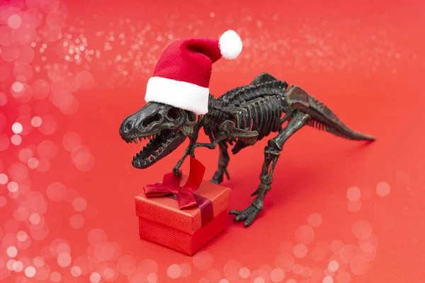 Noel Baba şapkalı dinozor iskeleti kırmızı arka planda duruyor. Yeni yıl hediyesiyle birlikte. Eski Yeni Yıl konsepti.