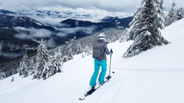 Genç kayakçı kayaklarıyla çakıllı bir yamaçta duruyor. Dağ yamaçları karla kaplı. Karla kaplı çam ağaçları