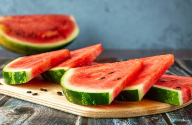 Tahta kesme tahtasının üzerinde üçgen şeklinde kırmızı karpuz parçaları var. Arka planda mavi bulanık bir arka plan ve yarım karpuz var..