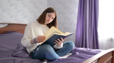 Genç güzel kız evde yatakta otururken bir kitap okuyor ve sayfaları karıştırıyor. Kopyalama alanı