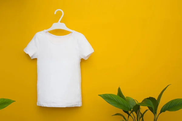 Camiseta blanca para niños colgada sobre un fondo amarillo junto a hojas verdes. Mocap. Espacio de copia —  Fotos de Stock