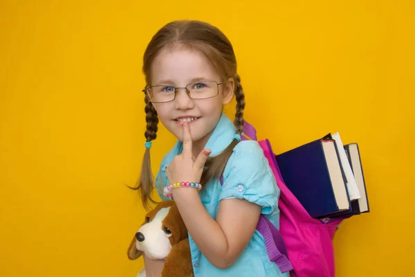 Wracać do szkoły. Szczęśliwa piękna dziewczyna w okularach z plecakiem szkolnym z książkami trzyma miękką zabawkę uśmiechniętą przemyślnie do kamery na żółtym tle. Wolne miejsce. Przestrzeń chroniona — Zdjęcie stockowe