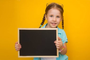 Okula dönelim. Saçları örgülü ve gözlüklü güzel küçük bir kız elinde sarı bir arka planda bir karatahta tutuyor..