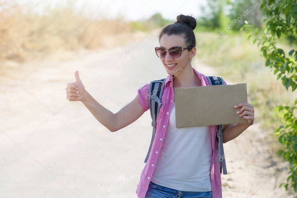 Beautiful cheerful young woman on the road waiting for a car and holding a cardboard with place for text. Hitchhiking during the summer holidays. Copyspace