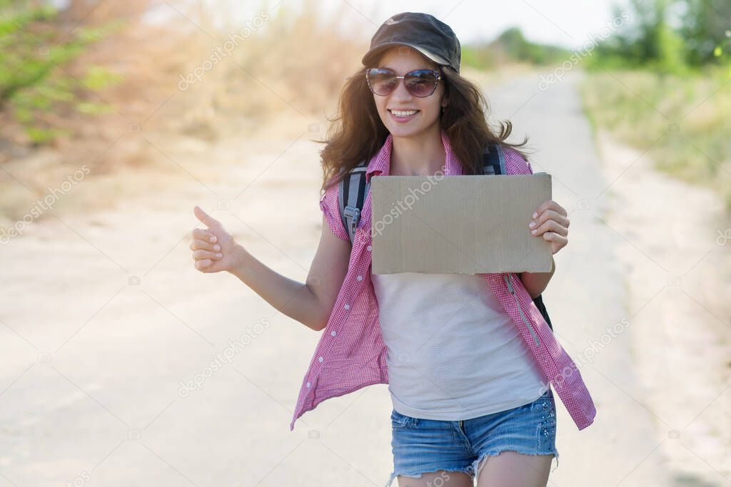 Hitchhiking woman travel. Beautiful young female hitchhiker in a cap and sunglasses smiles and catches a passing car by the road during a vacation, holds a cardboard with place for text and thumb up.