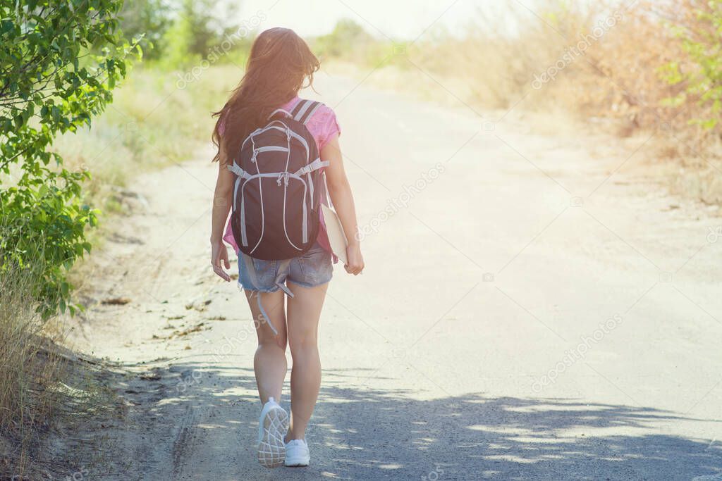 A young woman with long dark hair and a tourist backpack on her back is walking alone on the road while hitchhiking. Copyspace