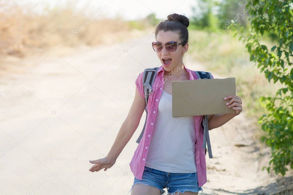 A girl hitchhiker with a backpack and sunglasses is surprised while standing on the road with a cardboard in her hands while hitchhiking. Copyspace