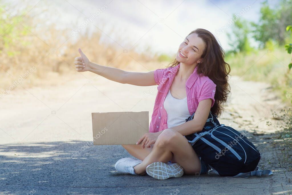 Happy young woman with a backpack and cardboard joyfully squints her eyes while sitting on the road and catches a passing car raising her thumb up. Hitchhiking during the summer holidays. Copyspace