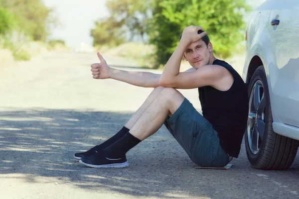 Handsome young sad man sits near his broken car and holds up his thumb up to stop a passing car. Copyspace