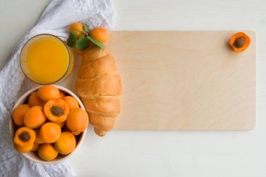Bir bardak taze meyve suyuyla olgun kayısılar ve kahvaltıda ahşap bir tahtada beyaz bir arka planda uzanan gevrek bir kruvasan. Temiz yeme konsepti, yakın çekim, telif alanı, düz yatak.