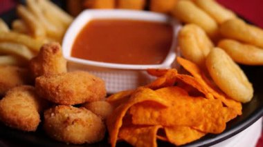 Yavaş çekimde bir tabak patates kızartması, nachos, kama, nugget ve balık kroketi ortada bir kap sosla siyah bir tabakta.