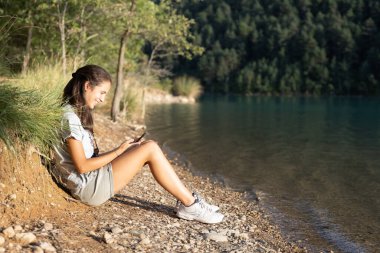 Öğleden sonra akıllı telefonunu kullanarak gölün kenarında oturan gülümseyen bir kız