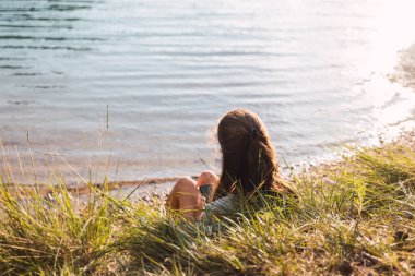 Arkadan bir kız cep telefonuyla gölün kenarında yatıyor.