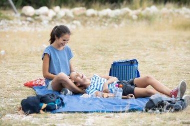 İki kız kardeş dağda konuşuyor ve gülüyor ve en küçüğü büyük kızın bacaklarında yatıyor.