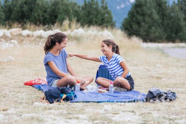 İki kız dağda çerez yerken yüksek sesle gülüyor ve küçük olan arkadaşlığın sembolü olarak yaşlının koluna dokunuyor.