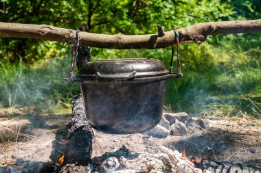 Açık havada yemek pişirmek için bir tencere çubuğa asılı. Odunların altı için için için yanar ve duman görünür. Arka plan bulanık. Saha mutfağı