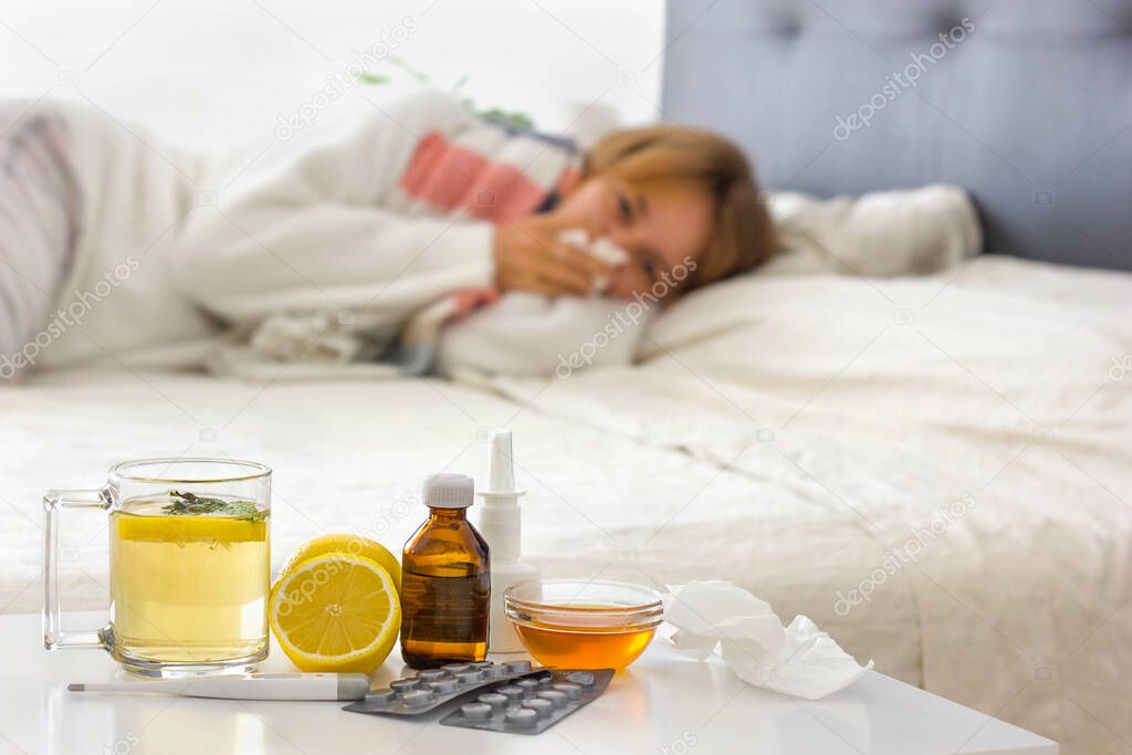 Selective focus on alternative medicine on the night table (hot drink, lemon, honey, pills, thermometer, nasal spray and crumpled handkerchiefs. Young sick woman laying on bed