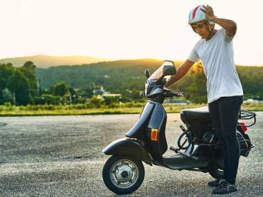 Kırık motorsikletli, endişeli ve arka planda günbatımı olan genç bir adam. Siyah saçlı, beyaz kasklı, İtalya bayrağı taşıyan beyaz tişörtlü ve dağlı siyah motosikletli bir çocuk.