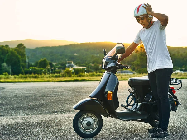 Kırık motorsikletli, endişeli ve arka planda günbatımı olan genç bir adam. Siyah saçlı, beyaz kasklı, İtalya bayrağı taşıyan beyaz tişörtlü ve dağlı siyah motosikletli bir çocuk.