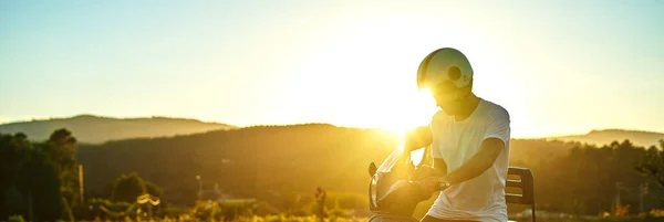 Arka planda günbatımı olan motosikletli çocuk panaromisi. Siyah saçlı, beyaz kasklı, İtalya bayrağı taşıyan beyaz tişörtlü ve arkasında dağlar olan siyah motosikletli bir çocuk..