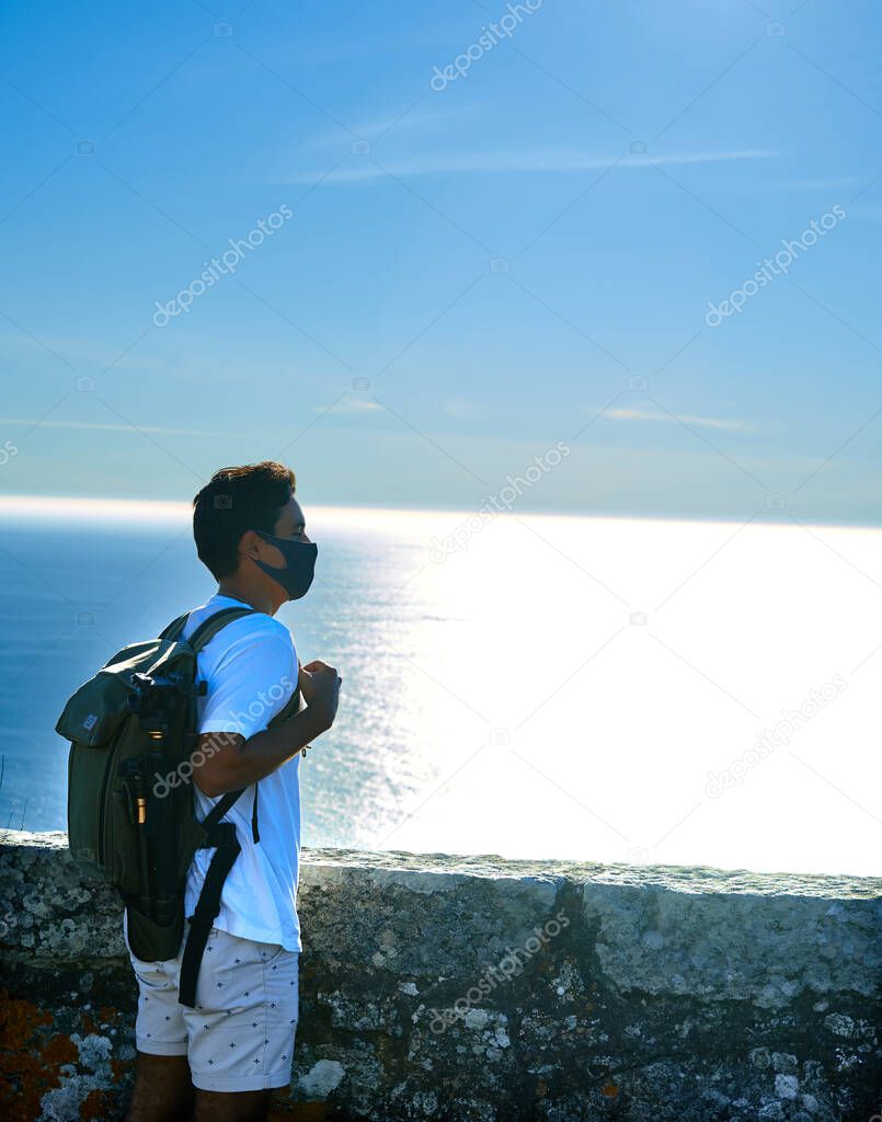 Young brunette traveler dressed in a white T-shirt with a photographic backpack and tripod watching the sea on the horizon. Traveler brunette teenager watching the sea from a viewpoint.