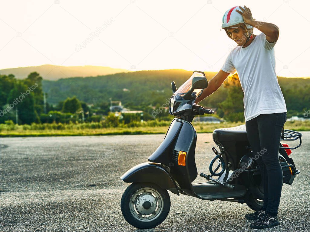 Young guy with broken motorcycle and looking worried and sunset in the background. Dark haired boy wearing white helmet with flag of Italy wearing white t-shirt and black motorcycle with mountains