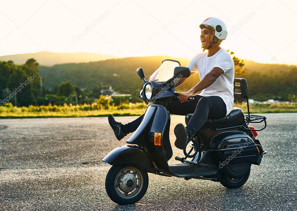 Boy having fun on a motorcycle with his feet raised and sunset in the background. Dark-haired boy with white helmet with flag of Italy with white t-shirt and black motorcycle with mountains