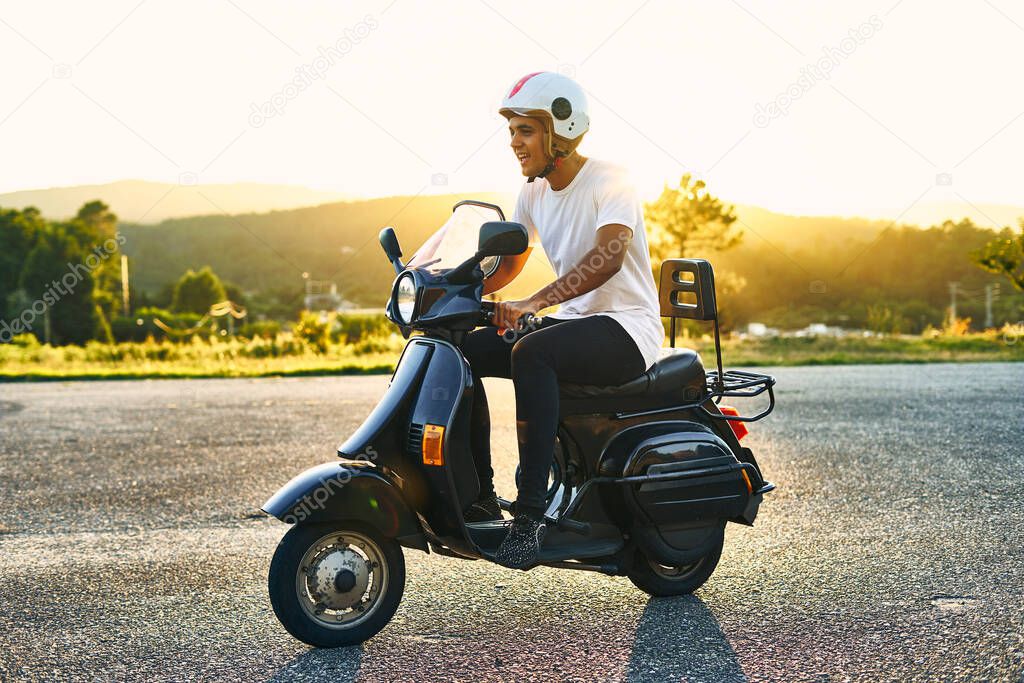 Boy on motorcycle with sunset in the background. Dark-haired boy with white helmet with flag of Italy with white t-shirt and black motorcycle with mountains in the background.