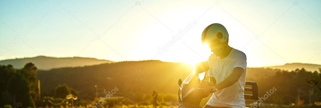 Panoramic of boy on motorcycle with sunset in the background. Dark-haired boy with white helmet with flag of Italy with white t-shirt and black motorcycle with mountains in the background.