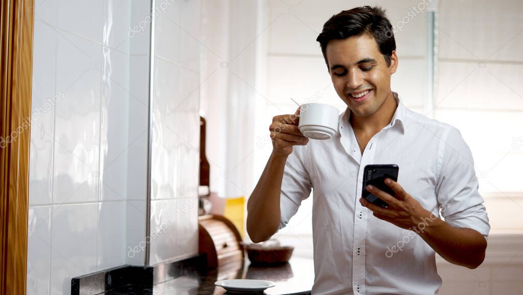 boy drinking coffee and watching his mobile. Man drinking coffee alone and watching social media on his phone while laughing. Teenager in white shirt in kitchen. Person laughing while having tea