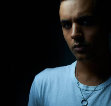 Siyah arka planda beyaz tişörtlü, siyah saçlı çocuk kameraya agresif bir şekilde bakıyor. Genç, kameraya ciddi bir bakış ve düşük ışıkla bakıyor. Gümüş kolyeli bir adam ciddi bir ifadeyle