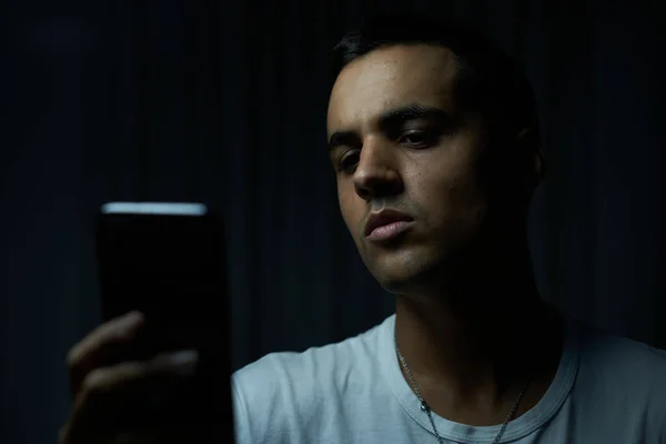 Siyah arka planda beyaz tişörtlü esmer çocuk cep telefonuna bakıyor. Ergen, ciddi gözleri ve düşük ışıkla telefona bakıyor. Gümüş kolyeli bir adam ciddi bir ifadeyle