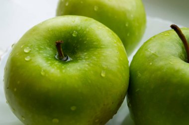 Üç olgun yeşil elma yakın planda, altın kenarlı beyaz bir tabakta yatıyor.