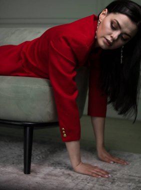 Beautiful young brunette girl in a red jacket lies on a gray armchair on a gray background