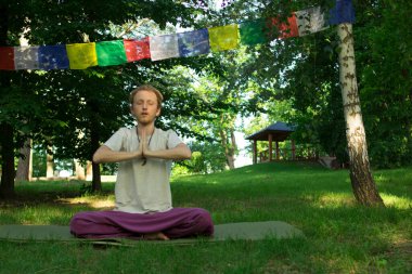 A man in a white T-shirt with red hair is meditating against the background of flags with Buddhist prayers