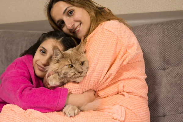 Beautiful mother and daughter in bathrobes are sitting on a gray sofa and holding a red cat in their arms. Good for as a billboard, flyer, wallpaper, poster