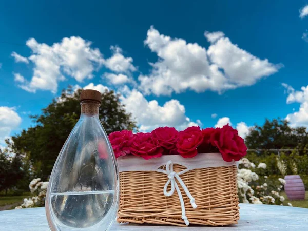 青い空に向かって灰色のテーブルの上に赤いバラとガラスの水のボトルを持つウィッカーライトブラウンバスケット — ストック写真