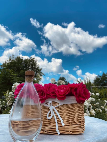 青い空に向かって灰色のテーブルの上には 赤いバラとバラのエッセンシャルオイルを持つガラスボトルとウィカーライトブラウンバスケット — ストック写真