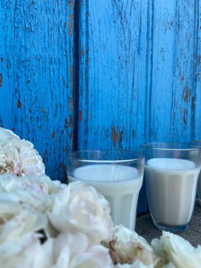 Yeşil yapraklı beyaz güllü şeffaf bardaklar, tahtadaki mavi ahşap yapının arka planında gri betonun üzerinde duruyor.