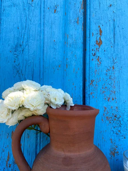 青い古い木の板の背景に古い茶色の粘土のジャグと白のバラ — ストック写真