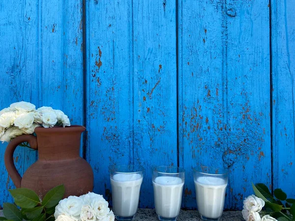木の板の青さを背景に 牛乳のグラスと白バラの古い茶色の粘土のジャグ — ストック写真