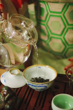 Yeşil kimonolu bir kız şeffaf bir çaydanlıktan ahşap tepside çayı olan beyaz bir kaseye su döküyor.